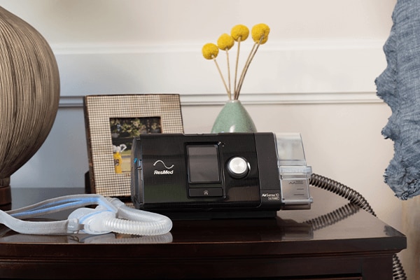 A ResMed CPAP machine and nasal mask on a bedside table, next to a lamp, a framed photo and some flowers.