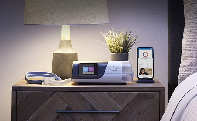 An Air11 sleep apnoea device, mask and a mobile phone showing myAir app on a bedside table.